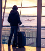 A person in an airport looking out the window as a plane takes off