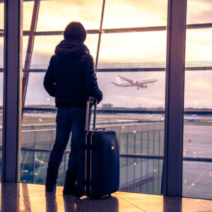 A person in an airport looking out the window as a plane takes off